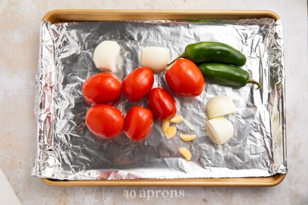 Roma tomatoes, jalapeños, garlic, and onions on a baking sheet lined with aluminum foil.