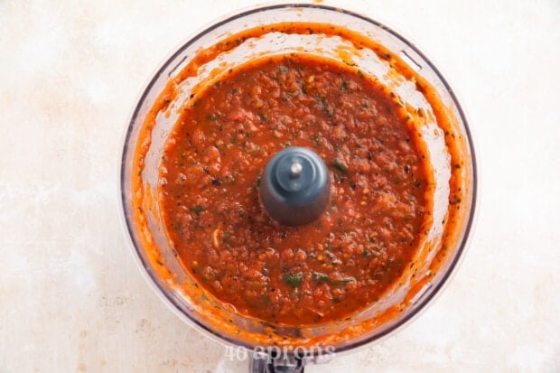 Overhead view of red roasted salsa in a food processor bowl on a light neutral countertop.