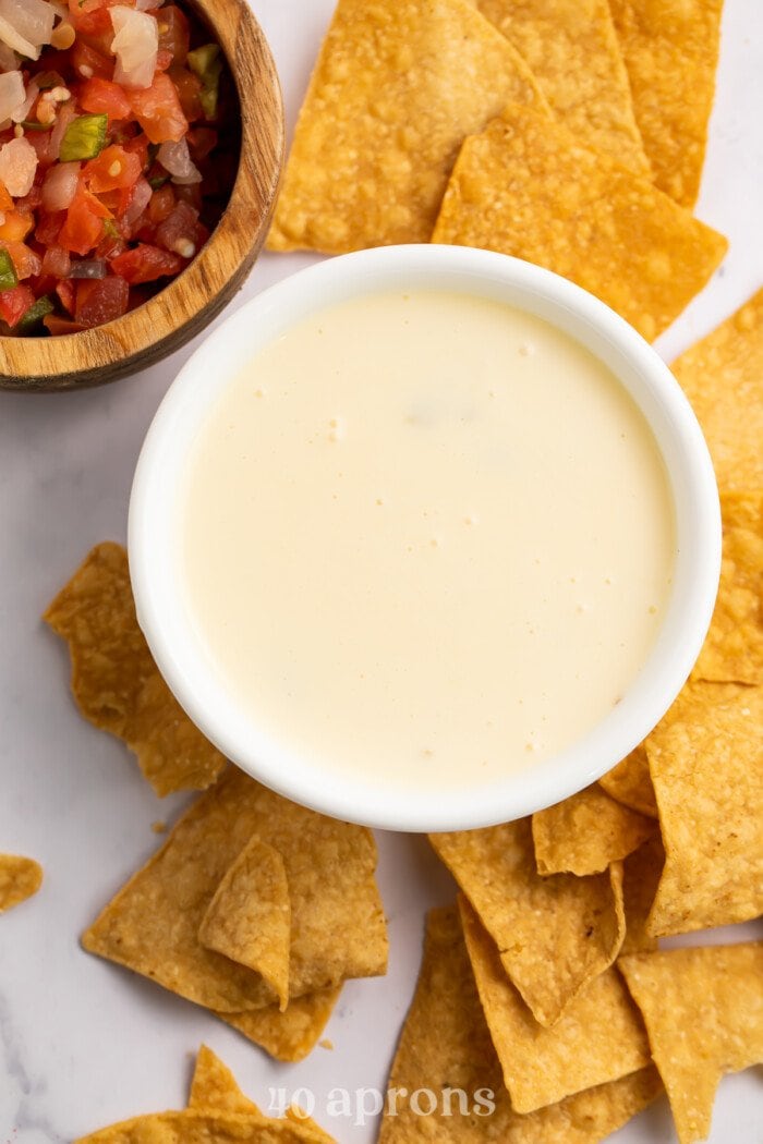 Overhead view of a bowl of white queso with a dollop of diced tomatoes in the center, surrounded by tortilla chips.