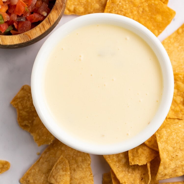 Overhead view of a bowl of white queso with a dollop of diced tomatoes in the center, surrounded by tortilla chips.