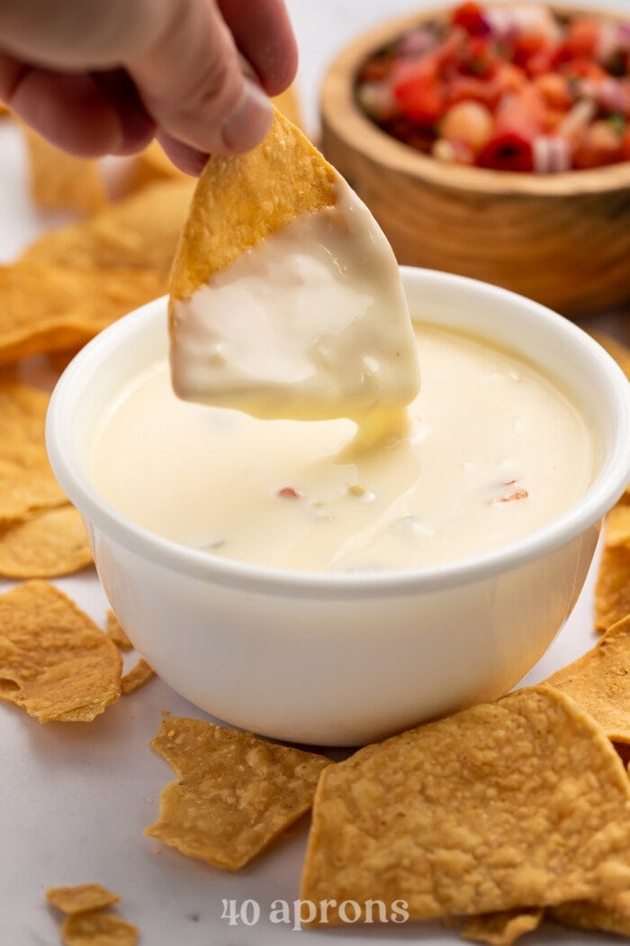 A tortilla chip that's been dipped in a bowl of white queso, hovering above the bowl and dripping queso.