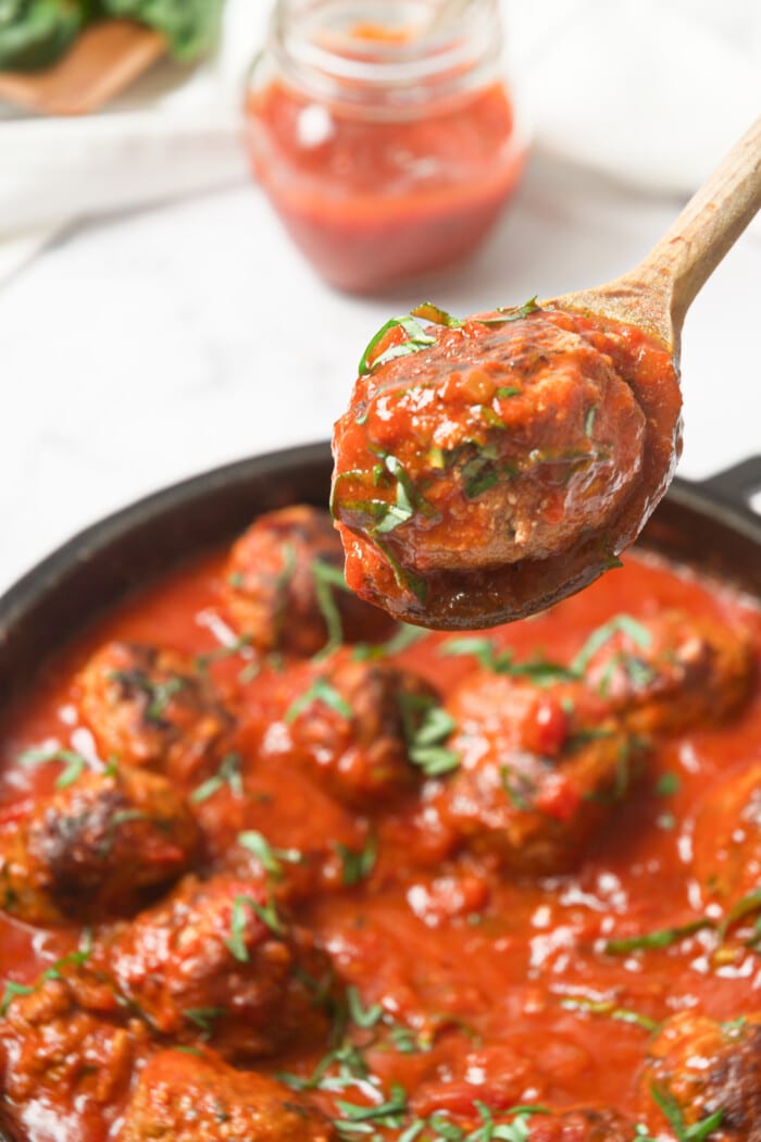 A Whole30 Italian meatball being lifted out of a cast iron skillet with a wooden spoon.
