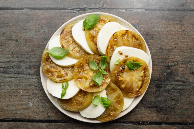 Overhead view of grilled green tomato slices alternating with mozzarella rounds on a plate with fresh basil.