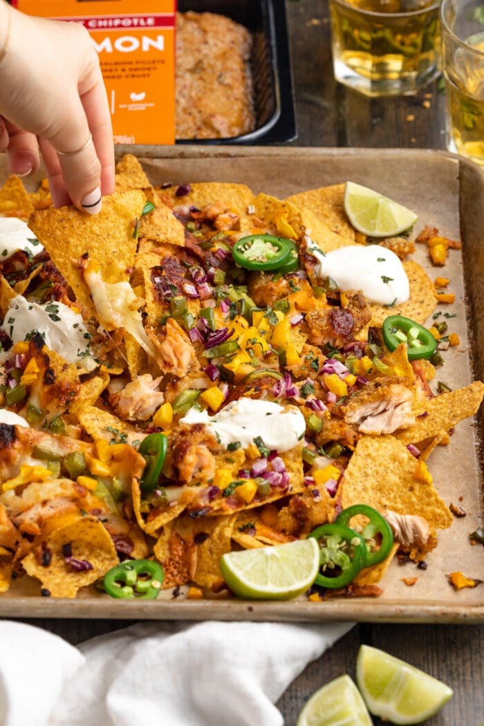 Angled view of a hand lifting up a tortilla chip with salmon from a baking sheet of salmon nachos.