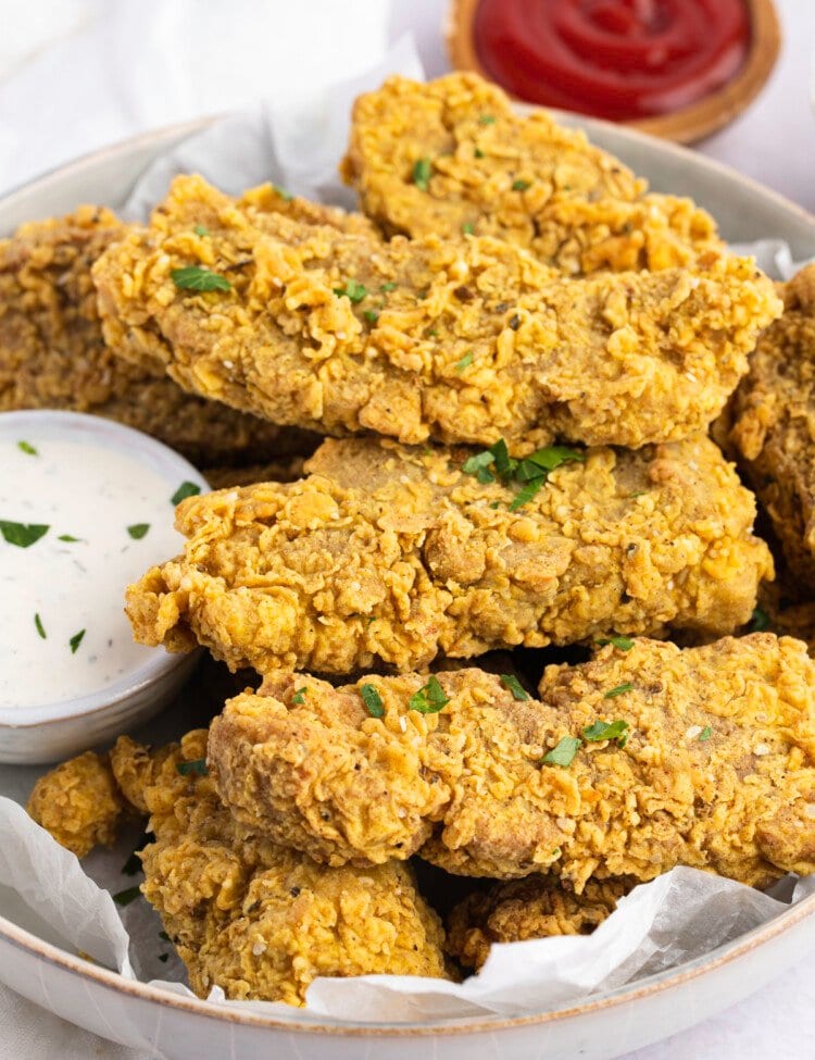 A pile of vegan seitan fried "chicken" tenders in a white bowl next to a small ramekin of ranch dressing with a small ramekin of ketchup in the background.