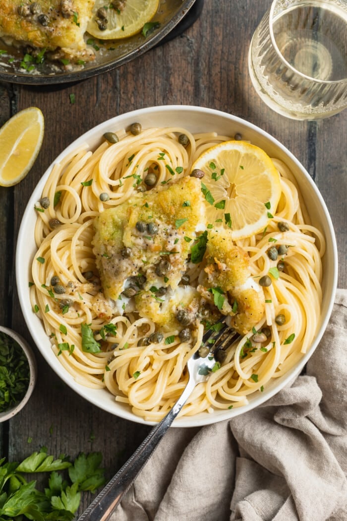 Overhead view of a bowl of potato-crusted cod piccata with spaghetti noodles, lemon slices, capers, and chopped parsley.