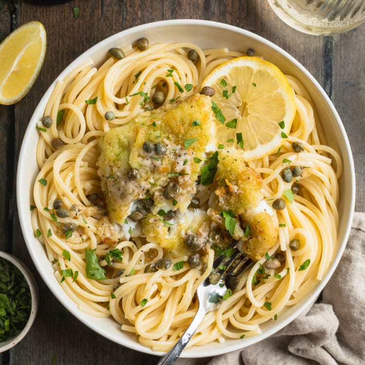 Overhead view of a bowl of potato-crusted cod piccata with spaghetti noodles, lemon slices, capers, and chopped parsley.