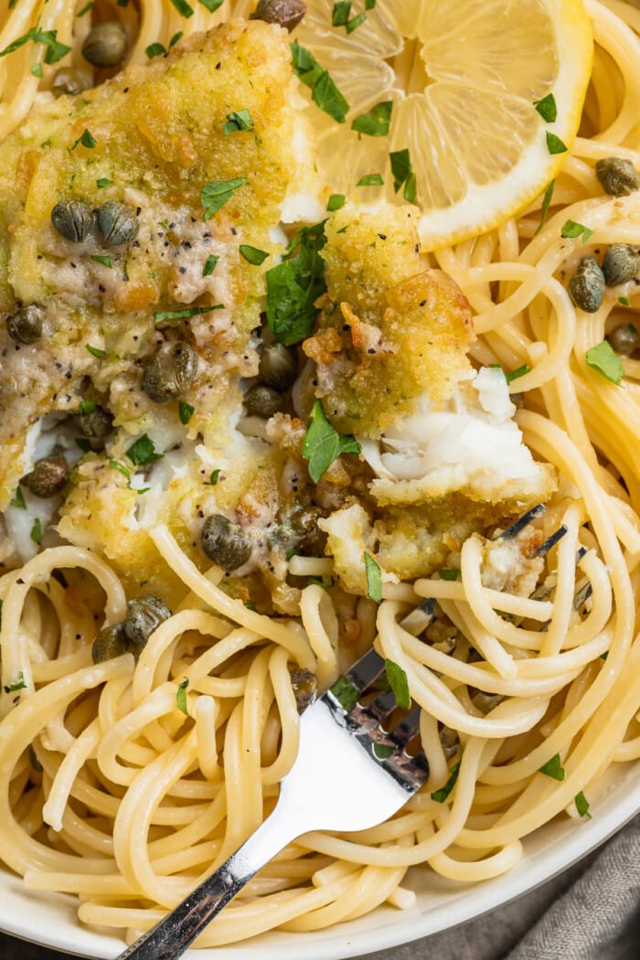 Close-up view of potato-crusted cod piccata with spaghetti noodles, lemon slices, capers, and fresh parsley.