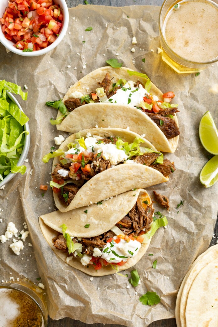 Overhead view of shredded beef tacos laying on a sheet of parchment paper, surrounded by toppings.