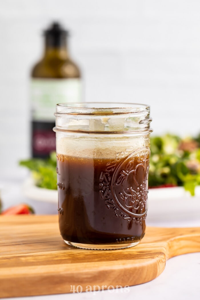 Side view of a glass jar of balsamic vinaigrette on a wooden cutting board with salad in the background.