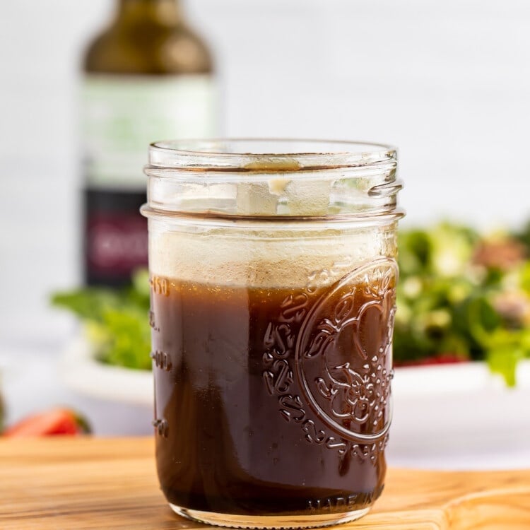 Side view of a glass jar of balsamic vinaigrette on a wooden cutting board with salad in the background.
