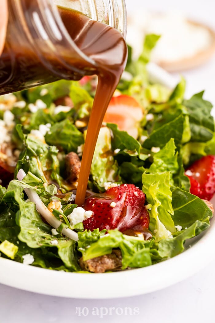 Balsamic vinaigrette pouring from a glass jar onto a large salad with green lettuce and red tomatoes.