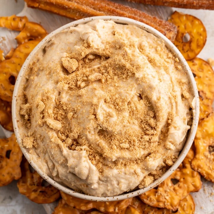 Overhead view of a small white bowl of churro dip surrounded by pretzel chips on a white countertop.