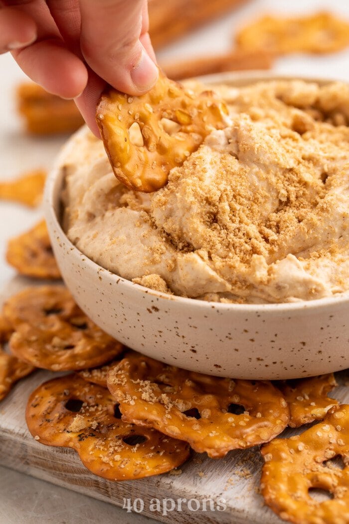 Side view of a hand scooping churro dip out of a small white bowl with a pretzel chip.