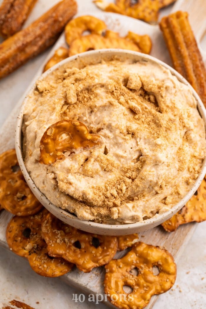 Angled view of a small white bowl of churro dip topped with crushed cereal surrounded by pretzels.