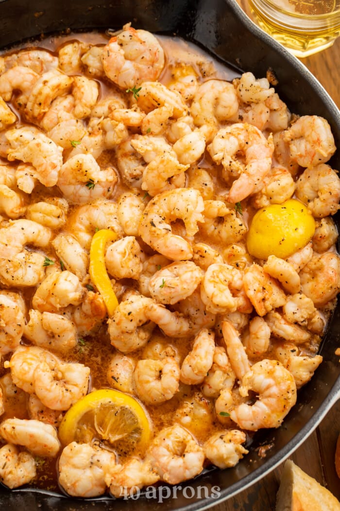 Overhead view of a large cast-iron skillet full of buttery BBQ shrimp with lemon circles.