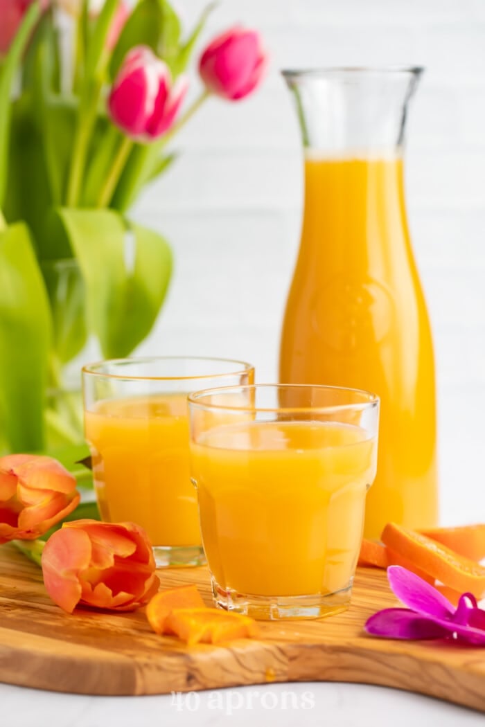 Side view of a glass of bright orange POG juice with a carafe of POG Juice and another glass in the background.