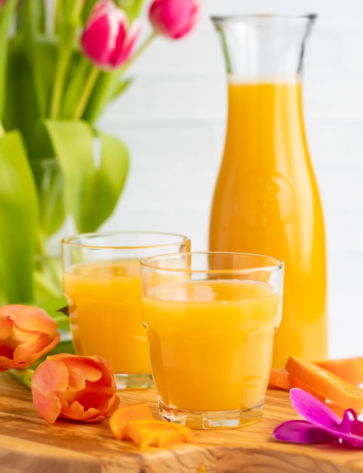 Side view of a glass of bright orange POG juice with a carafe of POG Juice and another glass in the background.