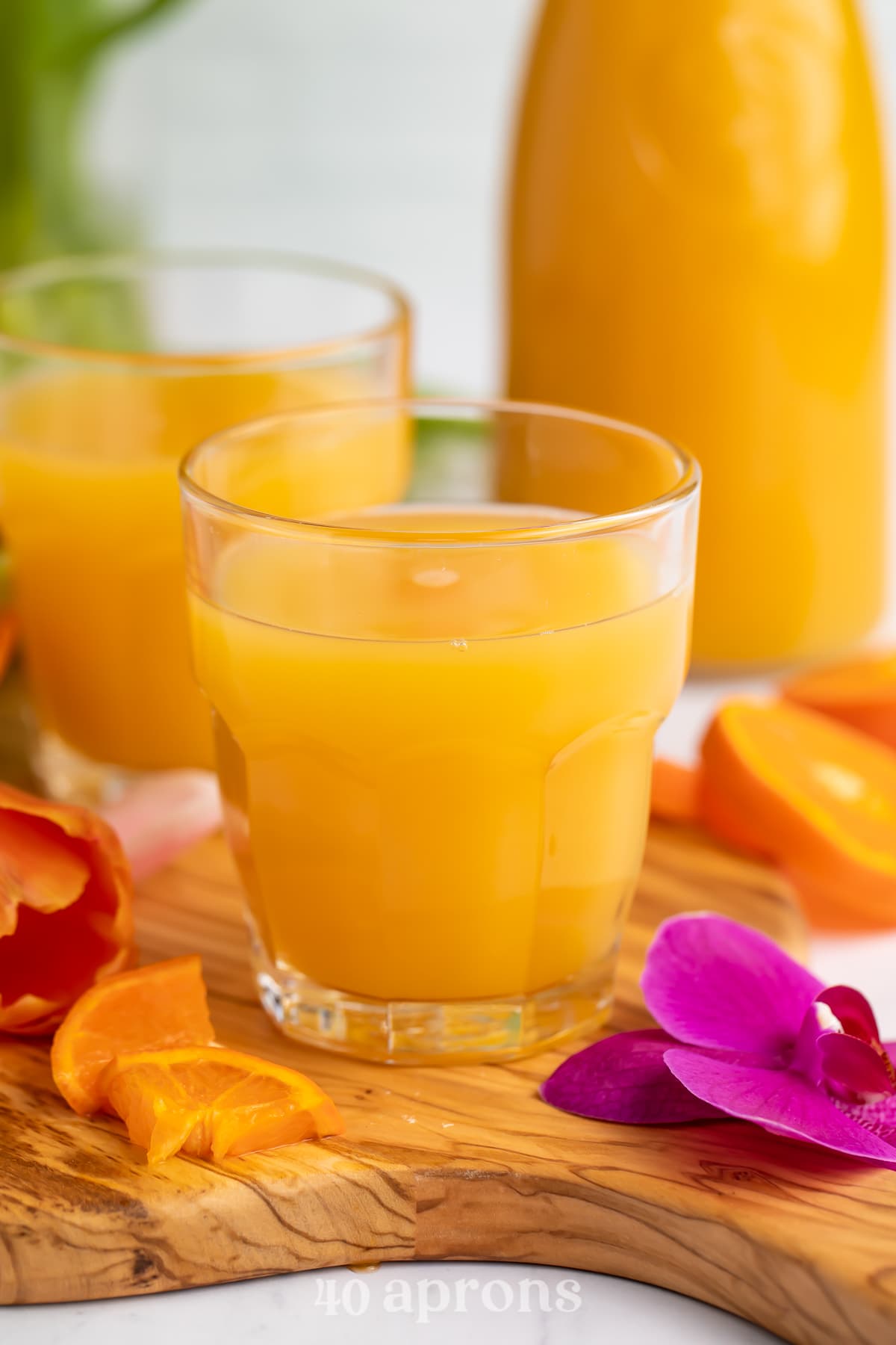 Side view of a glass of bright orange POG juice with a carafe of POG Juice and another glass in the background.