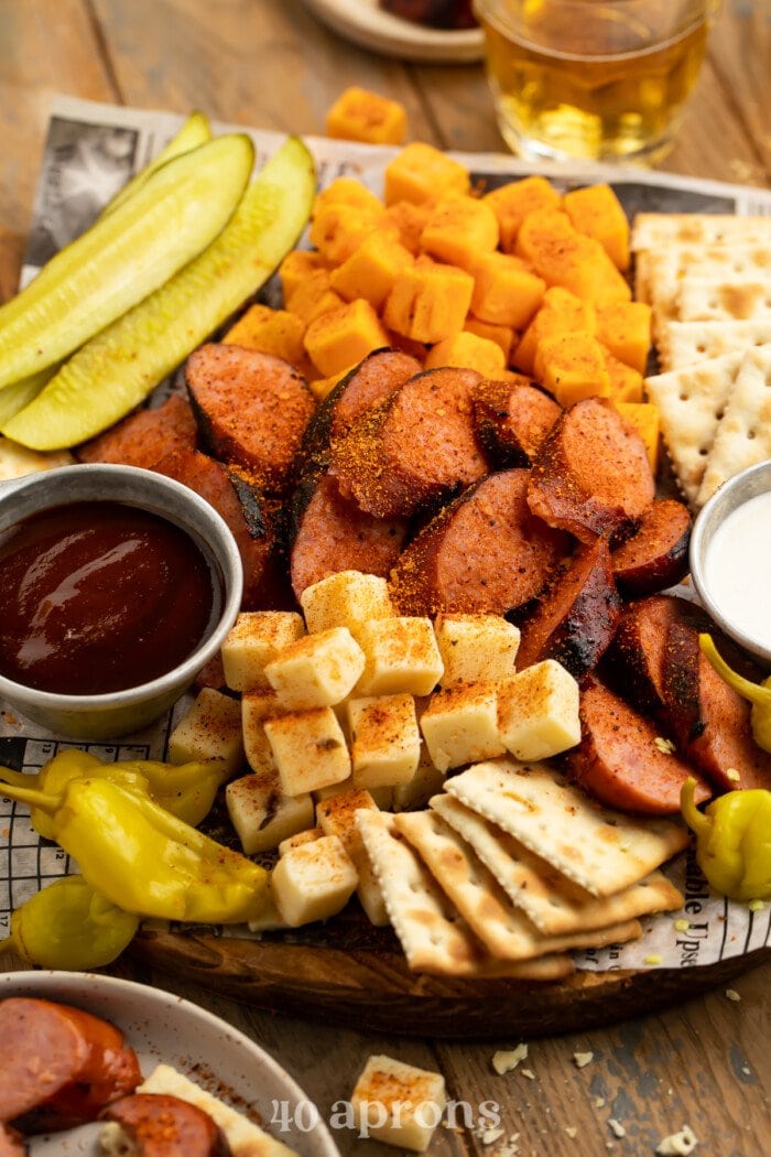 Close-up, overhead view of a Memphis-style cheese and sausage plate with pickles, crackers, ranch, and BBQ sauce on newspaper.