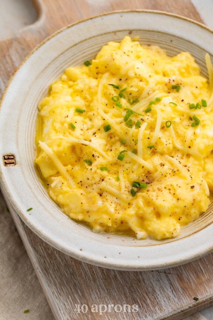 Close-up, overhead view of a bowl of truffled scrambled eggs garnished with chives on a wooden serving board.