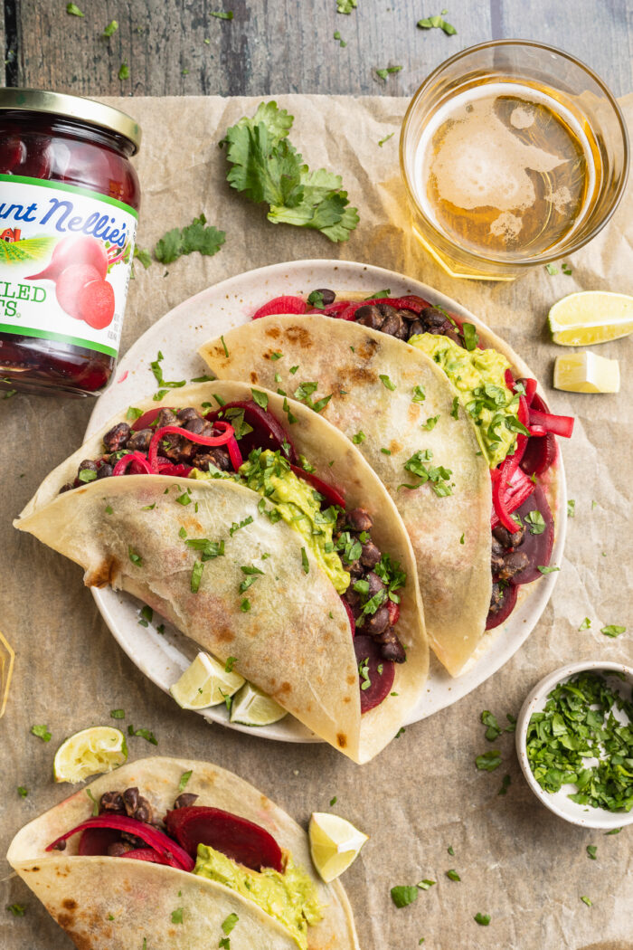 Overhead view of two pickled beet tacos on a plate surrounded by garnish and a jar of pickled beets.