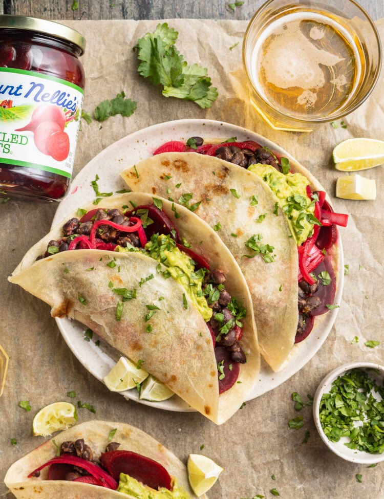 Overhead view of two pickled beet tacos on a plate surrounded by garnish and a jar of pickled beets.