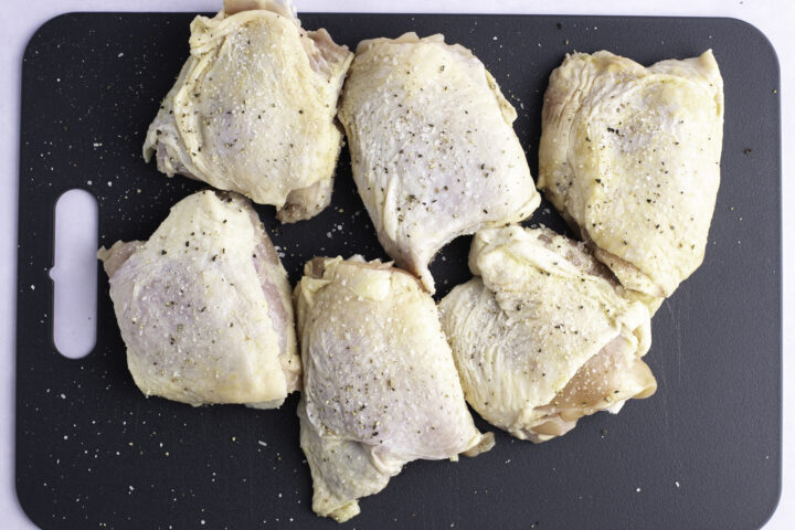 Overhead view of 6 raw chicken thighs seasoned with kosher salt and black pepper resting on a plastic black rectangular cutting board.