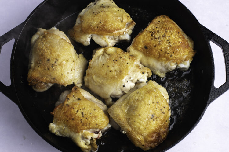 Overhead view of seared chicken thighs in a large black cast-iron skillet on a white table.