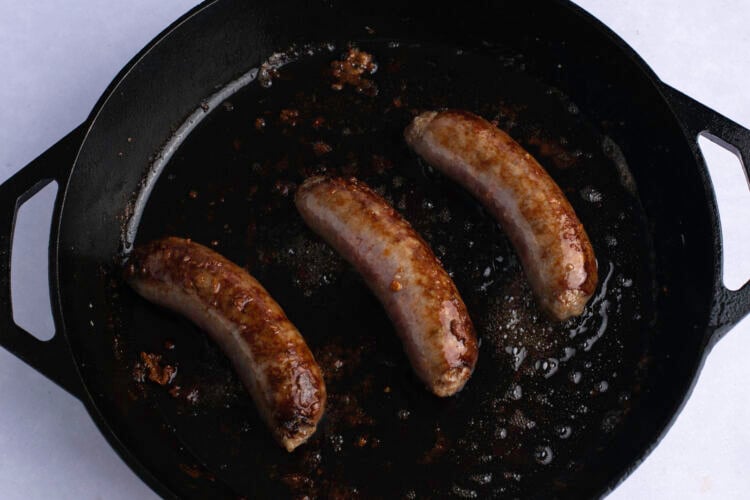 Overhead view of 3 Italian sausage links in a large black cast iron skillet on a white table.