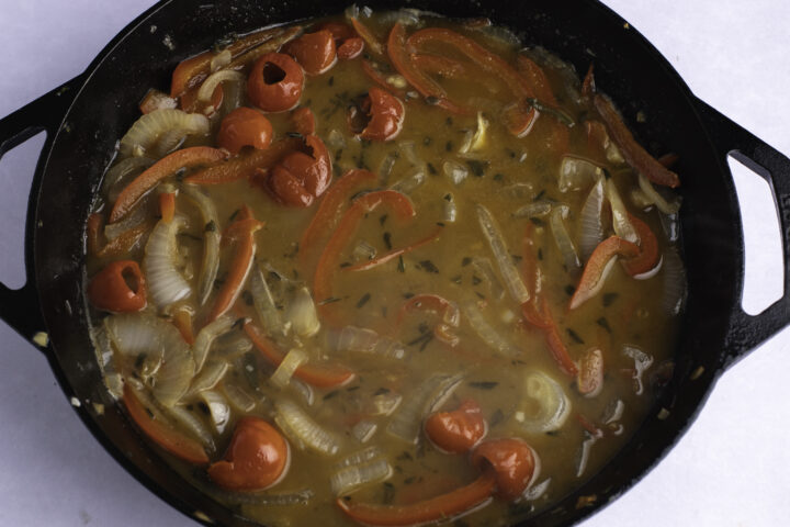 Overhead view of sauteed peppers, onions, fresh herbs, and white wine in a large cast iron skillet on a white table.