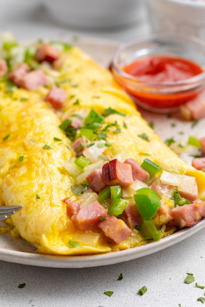 Angled view of a Denver omelette on a plate, with a bowl of ketchup in the background, and ham and green onions surrounding.