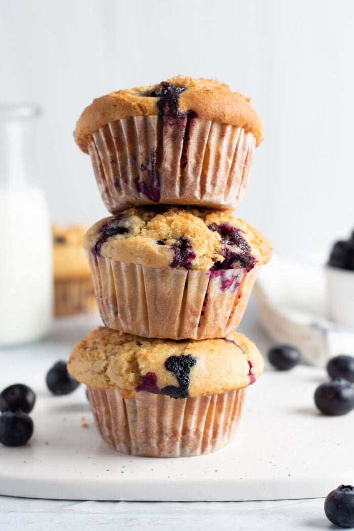 Side view of 3 gluten free blueberry muffins stacked on top of each other with stray blueberries on the counter.