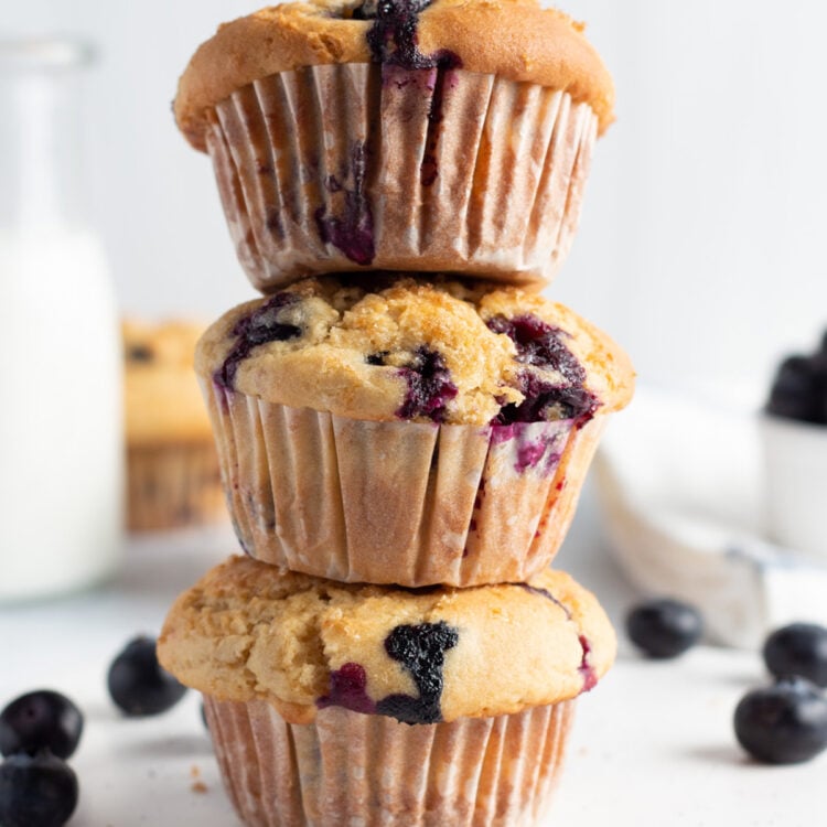 Side view of 3 gluten free blueberry muffins stacked on top of each other with stray blueberries on the counter.