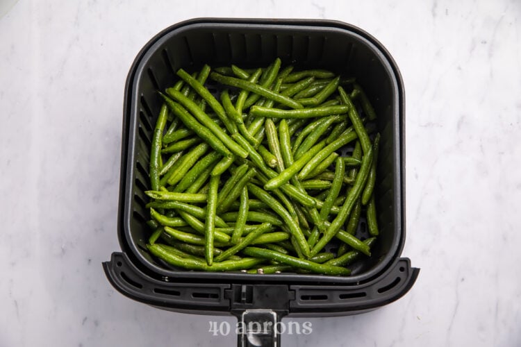 Overhead view of seasoned green beans in an air fryer basket on a white counter.