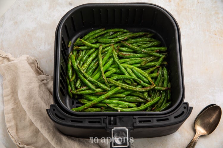 Overhead view of cooked green beans in an air fryer basket on a white counter.