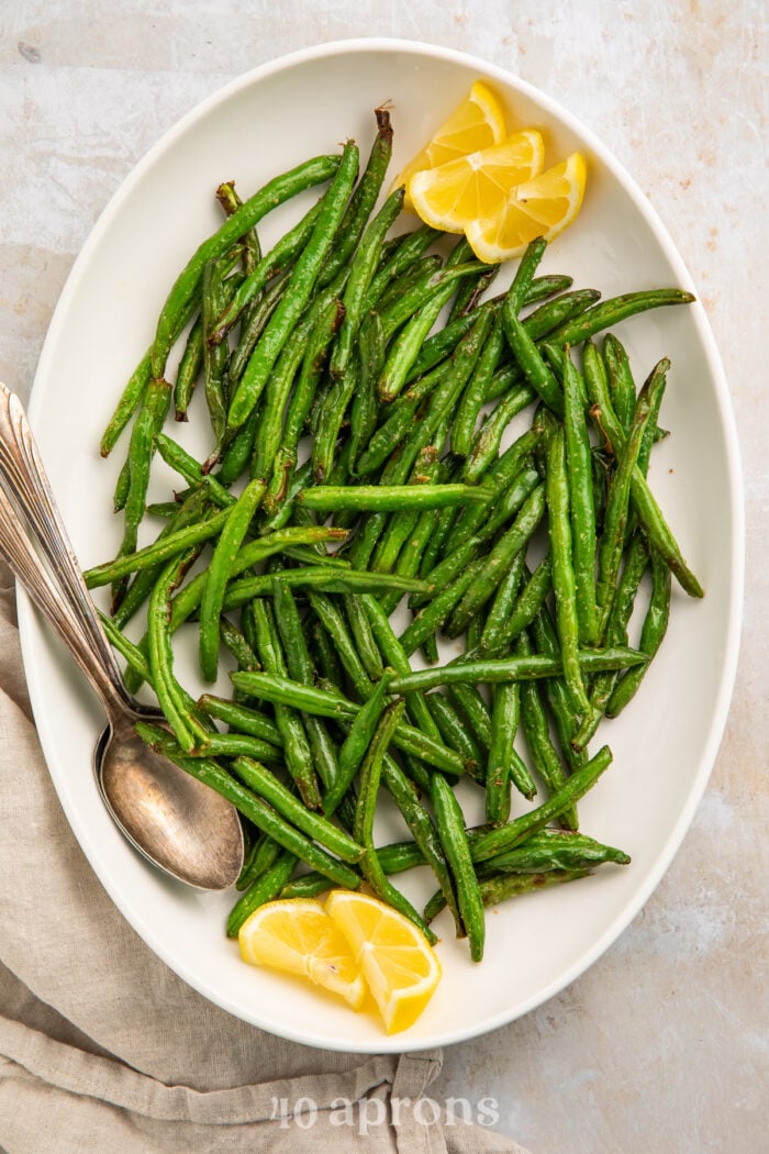 Overhead view of bright air fryer green beans on a white platter with lemon wedges and a silver spoon.