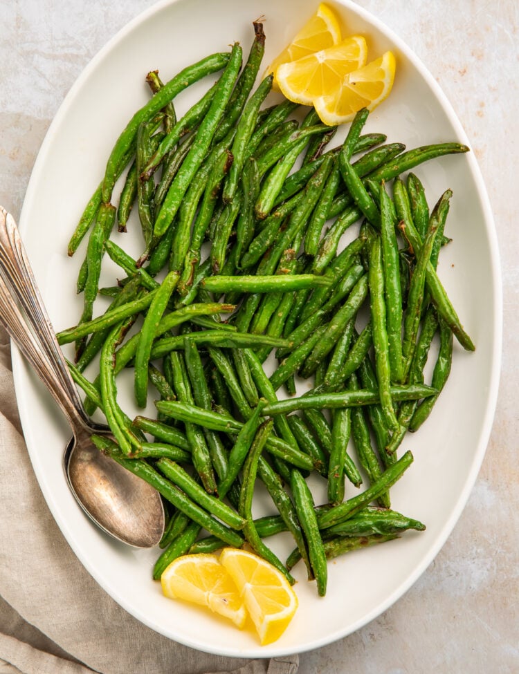 Overhead view of bright air fryer green beans on a white platter with lemon wedges and a silver spoon.