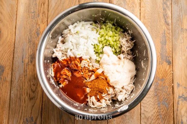 Overhead view of all ingredients for BBQ chicken salad in a large silver mixing bowl on a wooden counter.