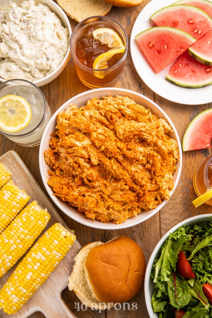 Overhead view of BBQ chicken salad in a bowl surrounded by corn on the cob, watermelon slices, and other barbecue essentials.