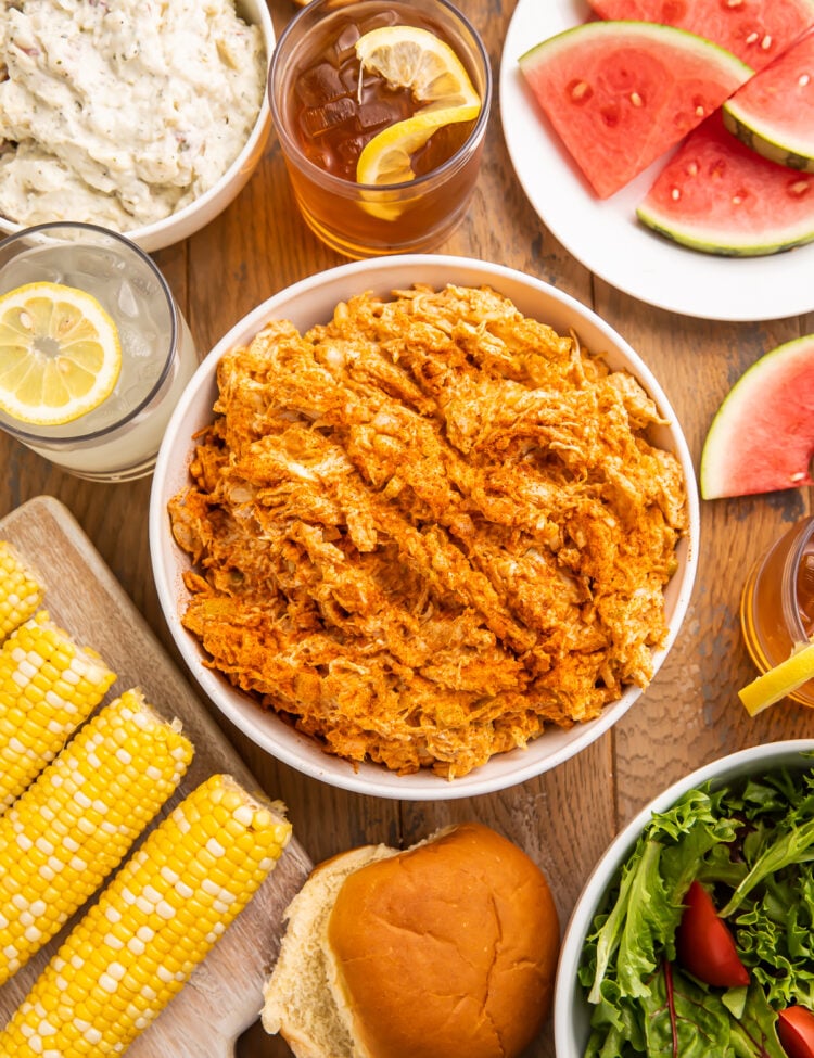Overhead view of BBQ chicken salad in a bowl surrounded by corn on the cob, watermelon slices, and other barbecue essentials.