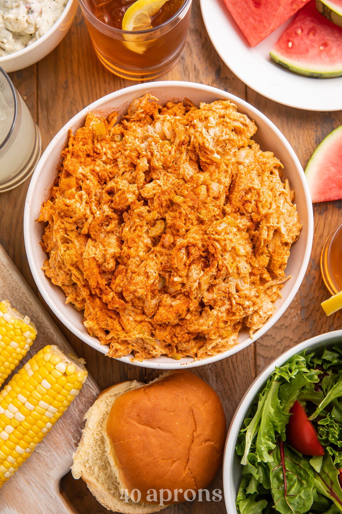 Overhead view of BBQ chicken salad in a bowl surrounded by corn on the cob, watermelon slices, and other barbecue essentials.