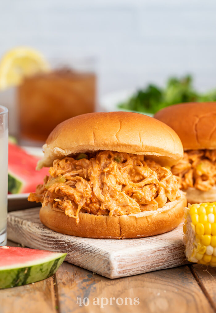 Side view of BBQ chicken salad on a burger bun with various barbecue essentials in the background.