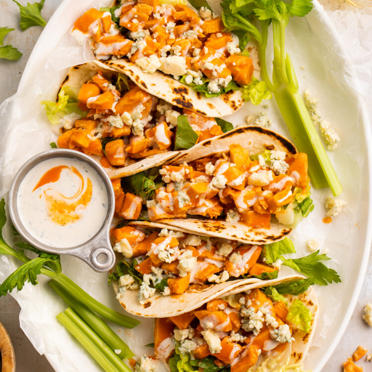 Overhead view of 5 buffalo chicken tacos lined vertically on an oval platter with celery sticks and ranch.
