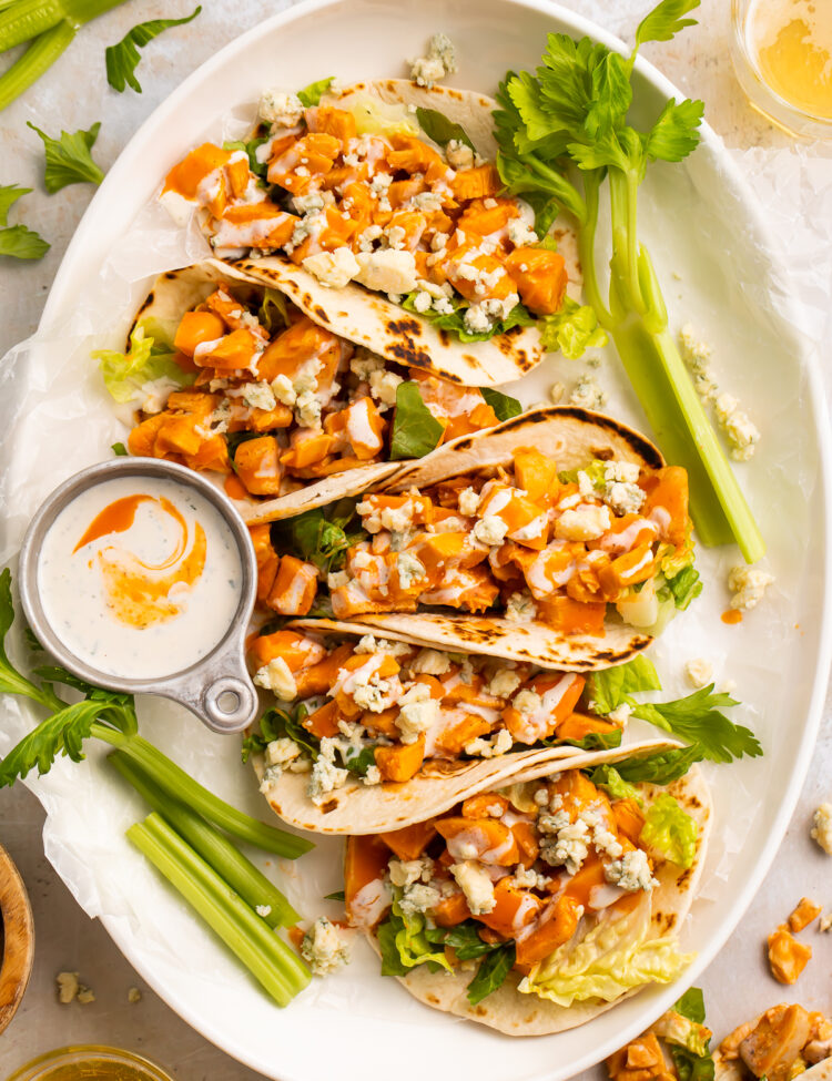 Overhead view of 5 buffalo chicken tacos lined vertically on an oval platter with celery sticks and ranch.