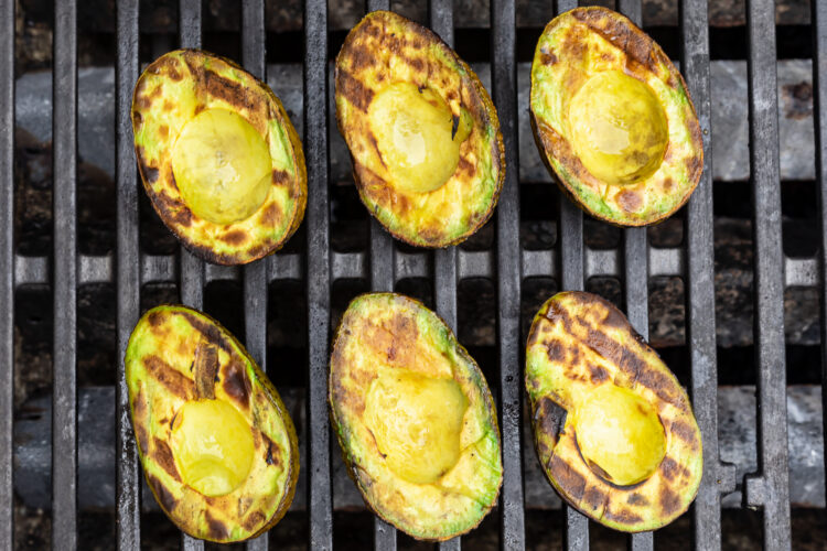 Overhead view of 6 avocado halves on a grill, with char marks criss-crossing on avocado meat.