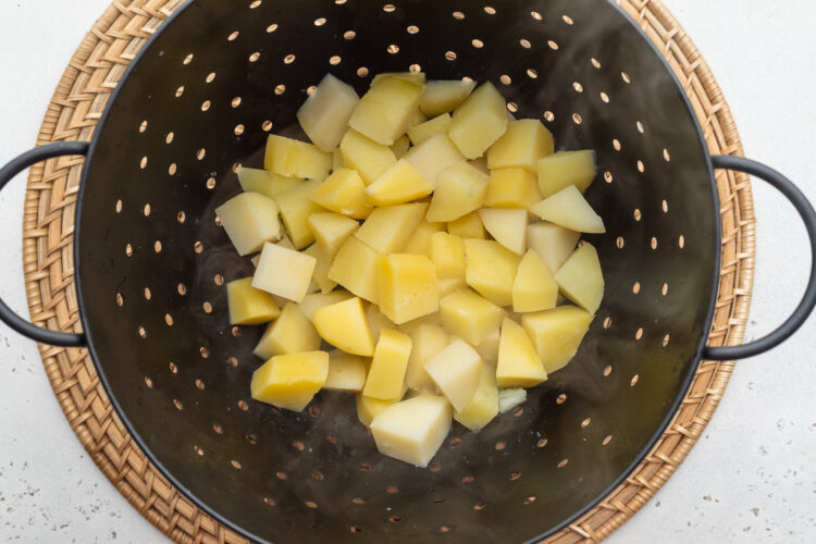Overhead view of boiled potato cubes in a large black colander on a neutral background.