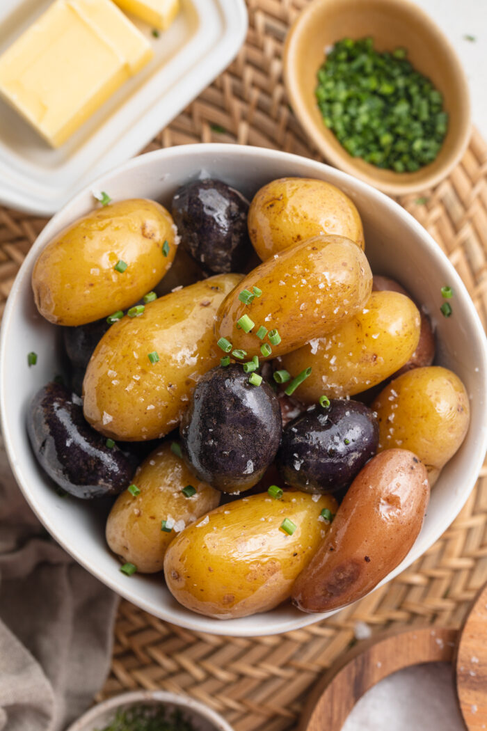 Overhead view of boiled light and dark potatoes in a white bowl on a wooden table surrounded by potato toppings.