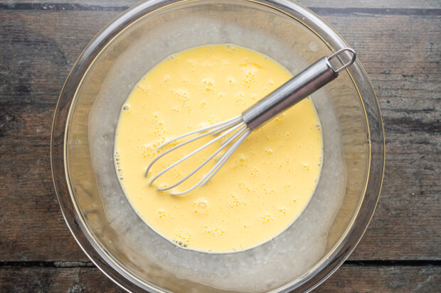 Overhead view of eggs and heavy cream whisked together in a large mixing bowl with a whisk.