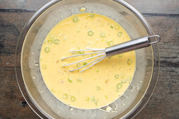 Keto quiche filling mixture in a large glass mixing bowl with a silver whisk on a neutral background.